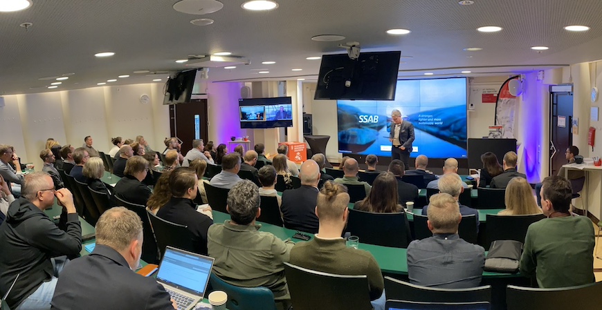 Fullsatt gradängsal med deltagare som sitter med ryggen mot kameran. Längst ner står föreläsare med en presentation som visas på en skärm med föreläsare online. Är ett hybridevent med deltagare på på plats och online.